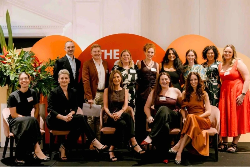 A diverse group of people pose smiling in front of a bold orange and red backdrop with plants on the side, suggesting a formal event or conference setting.