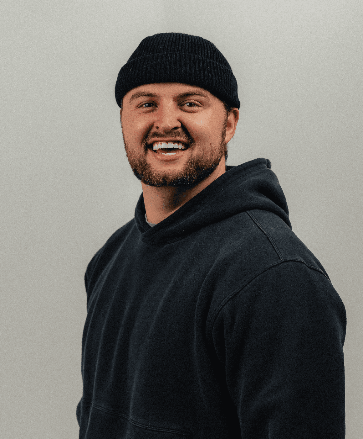 A man wearing glasses, a black hat, and a dark jacket looks confidently at the camera against a light background.