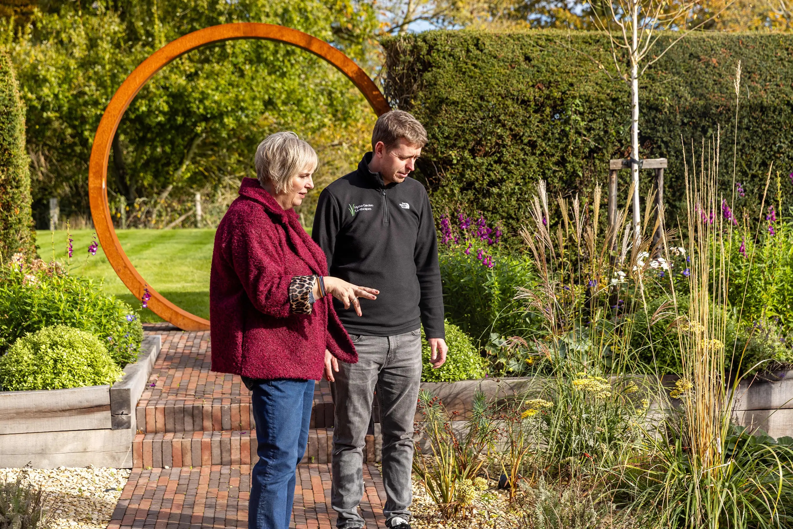 Two people converse in a garden, surrounded by plants and greenery, with a circular structure in the background.