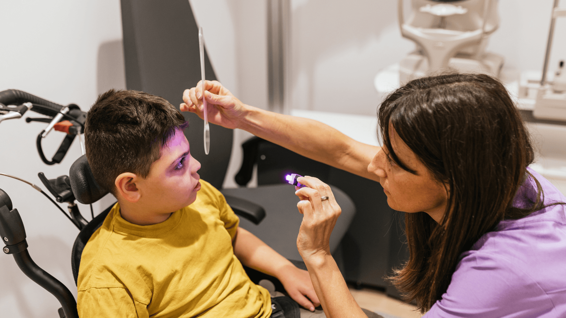 care professional examines a young child’s eyes using a light and vision tool in a clinical setting, while the child sits in a supportive chair during the exam.