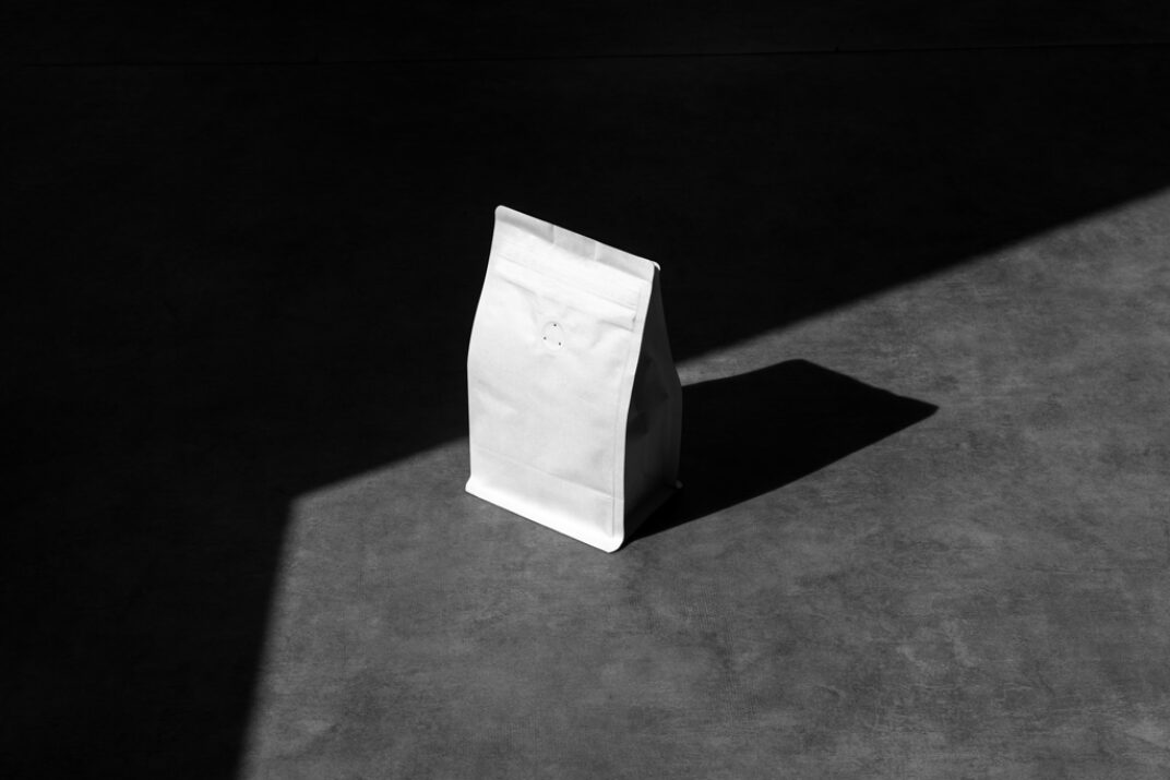 Minimalist paper bag mockup in stark black and white lighting. The bag stands upright on a concrete floor, with sharp shadows creating a dramatic contrast, perfect for branding designs.