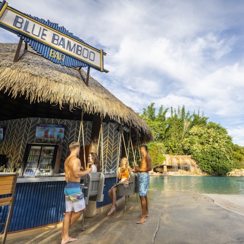 Трима души разговарят в бар със сламени покриви, наречен "Син Бамбу Бар", до басейн с зеленина на заден план.