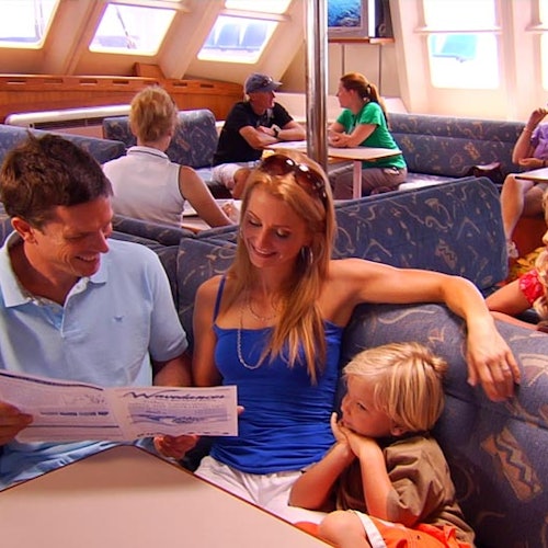 ครอบครัวหนึ่งนั่งอยู่ในพื้นที่เลานจ์ของเรือ ผู้ปกครองกำลังดูเมนู ในขณะที่เด็กนอนหนุนแขนแม่ของเขา ผู้โดยสารคนอื่น ๆ มองเห็นในพื้นหลัง