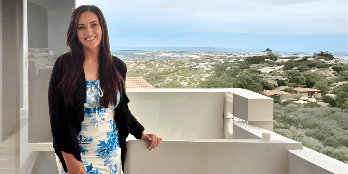 Nikita Lee Rindel standing on a balcony, smiling with a scenic view in the background.