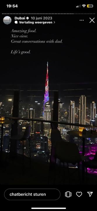 Smiling man with windswept red hair outdoors, wearing a black jacket and green backpack. Bright sunlight and a blurred natural background convey joy and adventure.