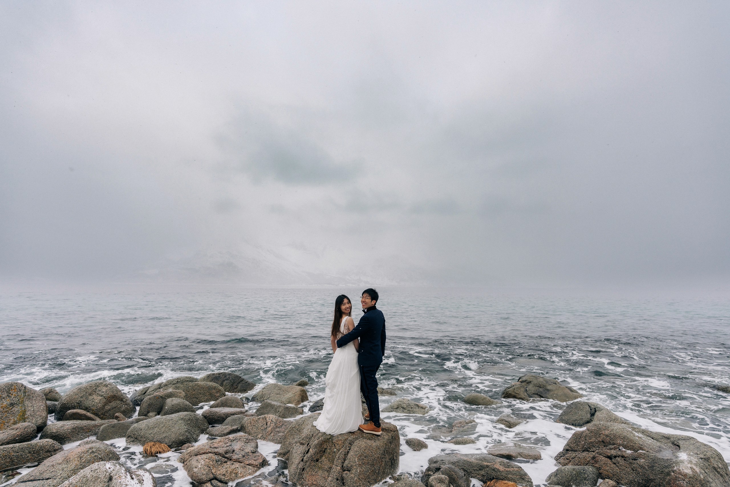 The couple standing together on dark coastal rocks, surrounded by pale surf and a fog-softened open sea.