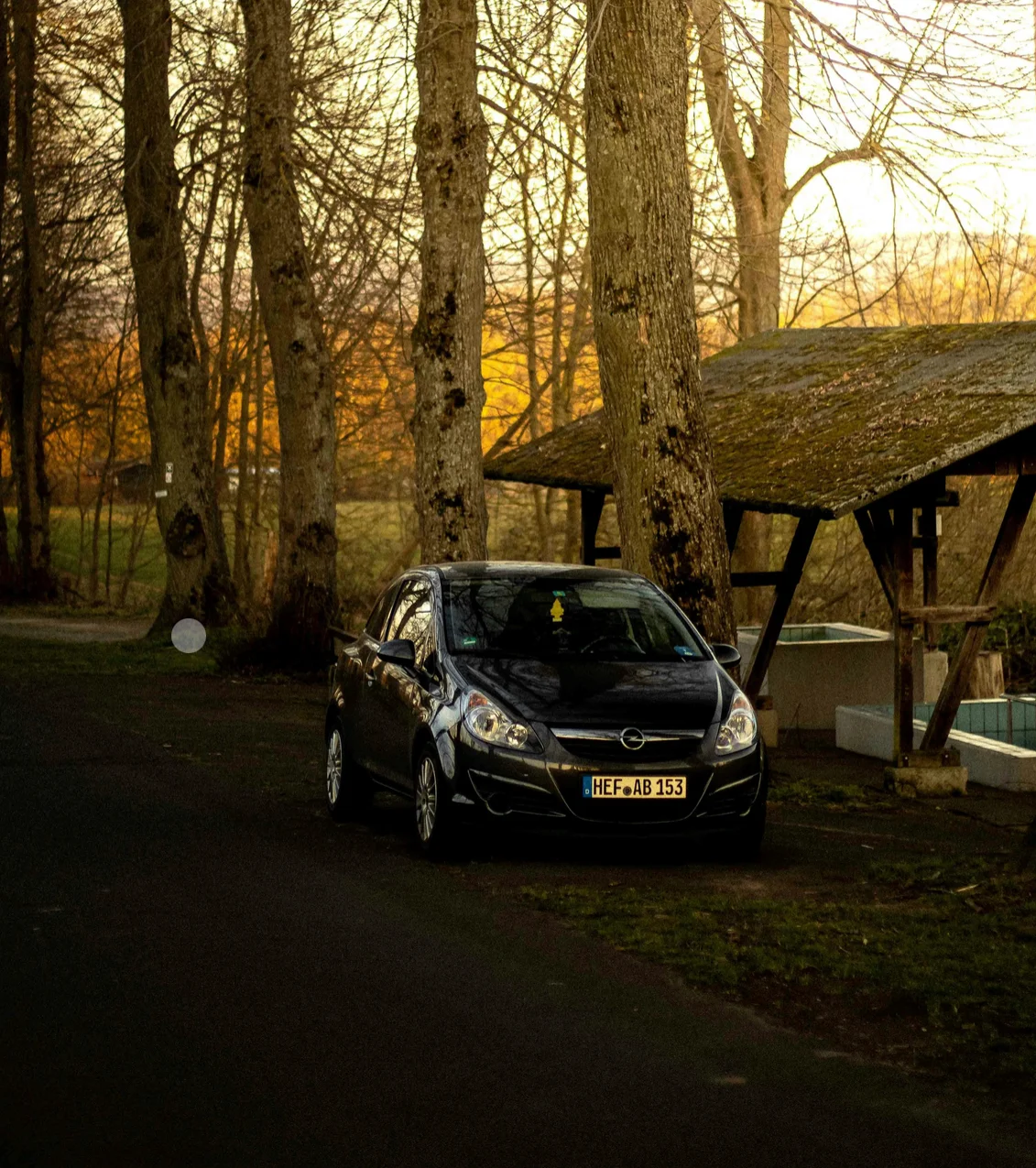 Une Opel Corsa stationnée dans une allée boisée au coucher du soleil. L’éclairage doré met en valeur les arbres nus et crée une atmosphère calme, où la petite citadine se fond dans un décor naturel et chaleureux.
