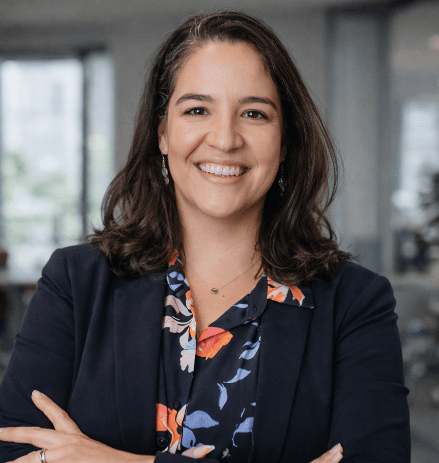 A smiling young woman with curly hair, wearing a purple shirt, stands in a bright indoor setting.
