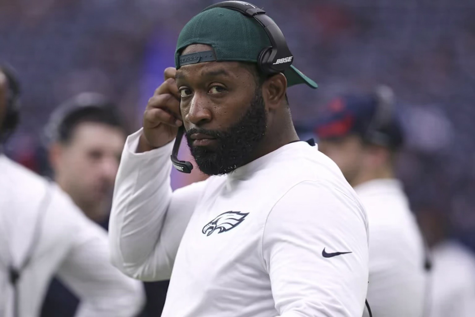 Allen Smith wearing a green cap, headset, and a white shirt with an Eagles logo stands on the sidelines, looking toward the camera with a focused expression.