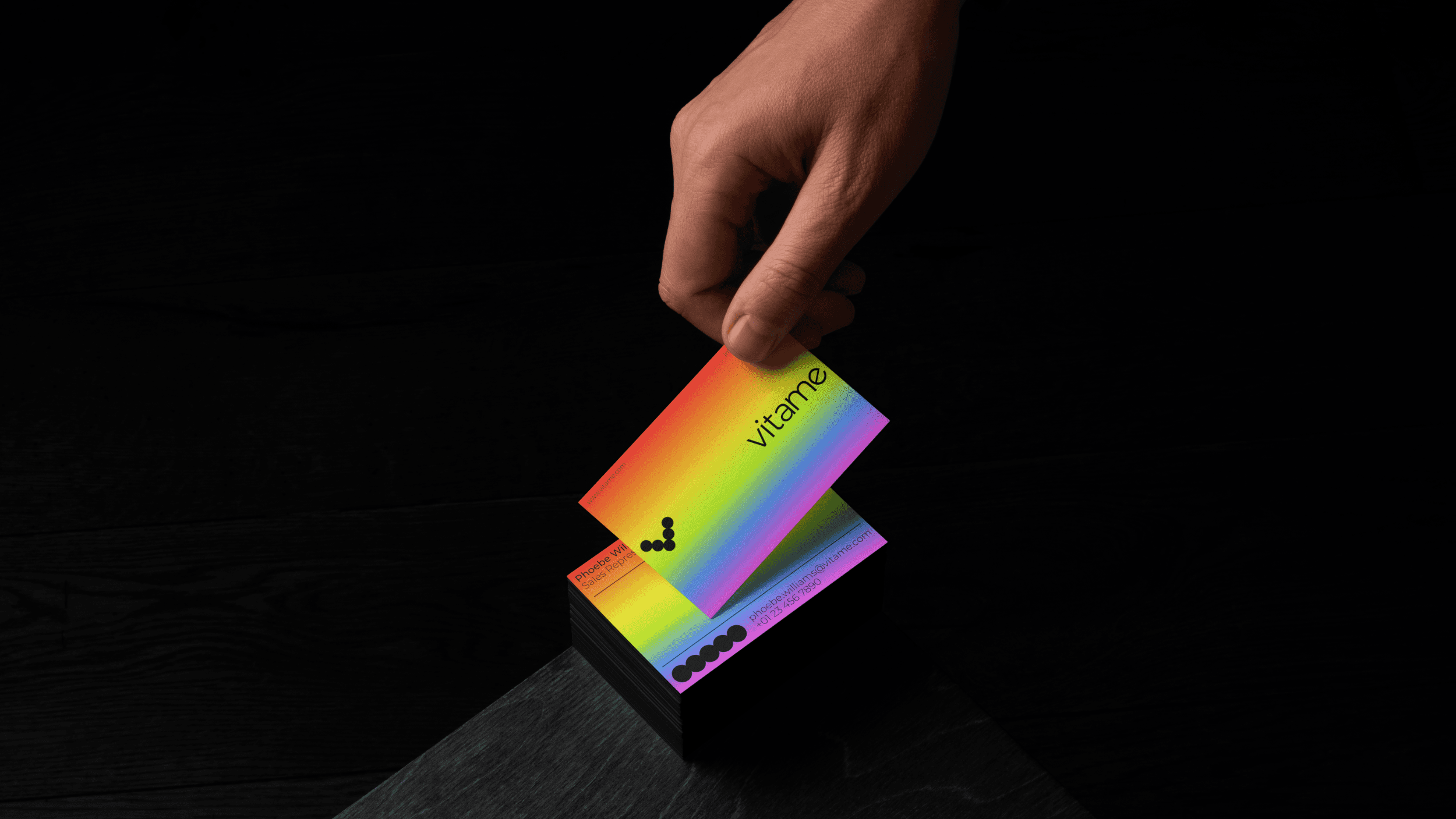 A stack of business cards sitting on the corner of a table. The business cards feature a vibrant rainbow gradient. A person's hand is reaching out to grab the card from the top of the stack.