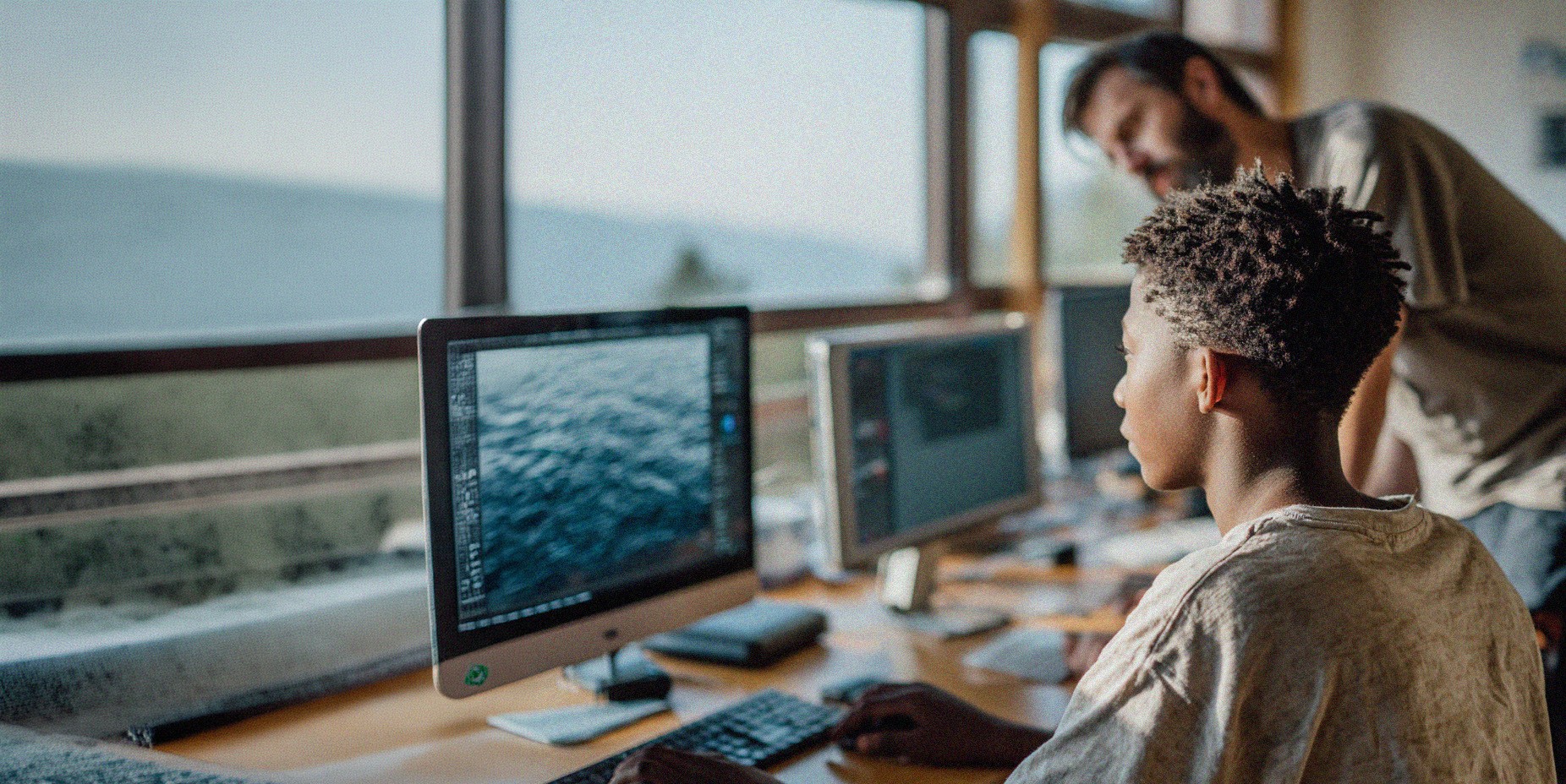 A young person with short, textured hair sits at a desk working on a desktop computer, focused on the screen. An adult stands slightly behind and to the side, leaning in as if offering guidance or instruction. The workspace includes multiple monitors, a keyboard, and other desk items, with large windows in the background letting in natural light, suggesting a classroom or learning environment.
