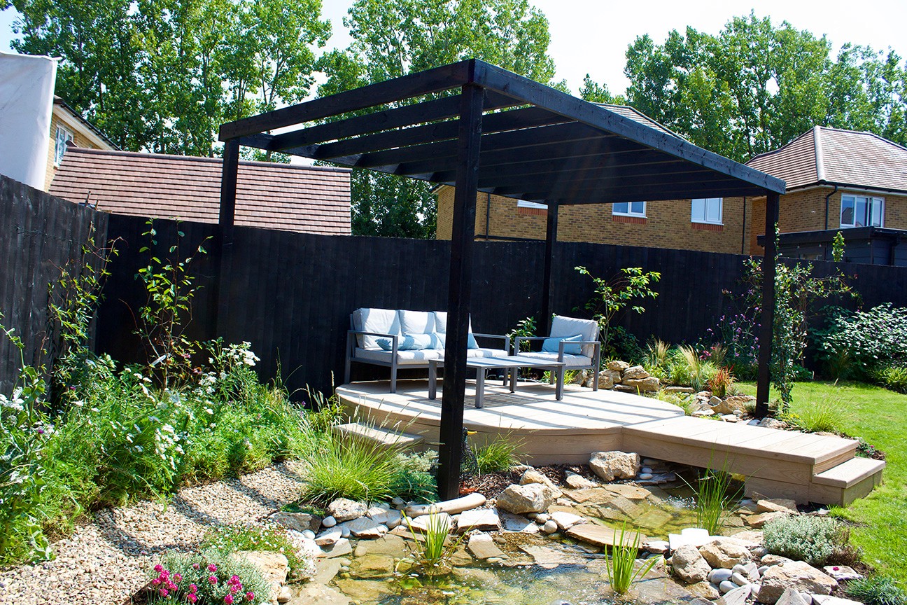 A backyard scene featuring a shaded seating area with a black pergola, surrounded by lush greenery and a pond.