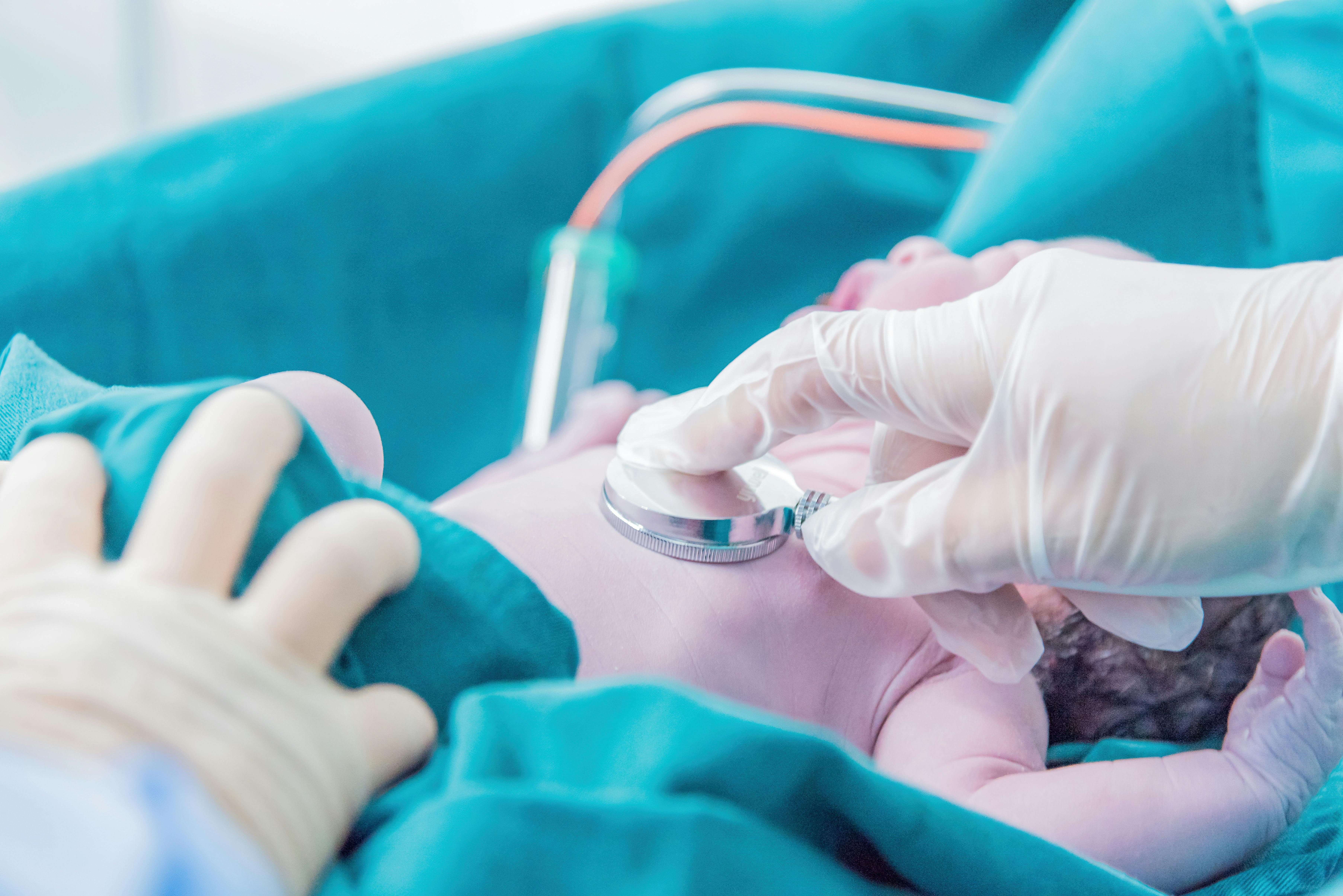 A newborn baby lies on blue medical cloth. A gloved hand holds a stethoscope to the baby's chest.