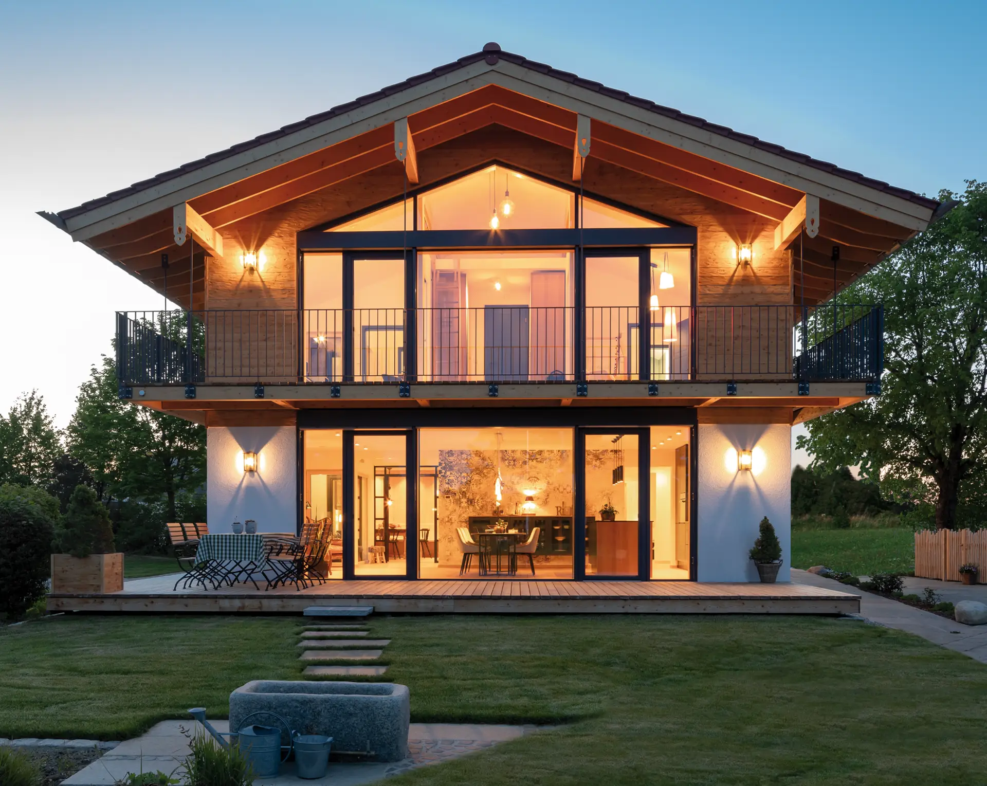 Modernes Holzhaus mit großzügiger Glasfront, Balkonen und Terrasse im Abendlicht, umgeben von gepflegtem Garten.