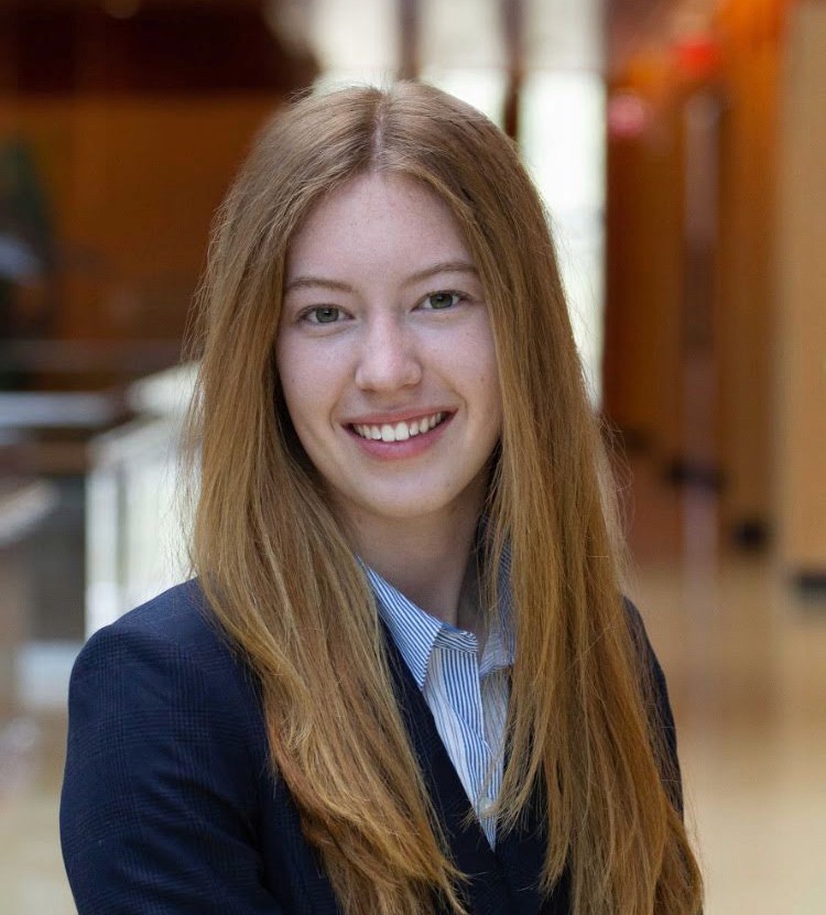 Smiling young woman with long hair, dressed in a blazer, standing in a well-lit indoor space.