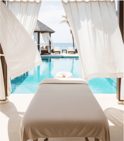 Poolside massage bed with flowing white curtains and ocean view on a sunny day.