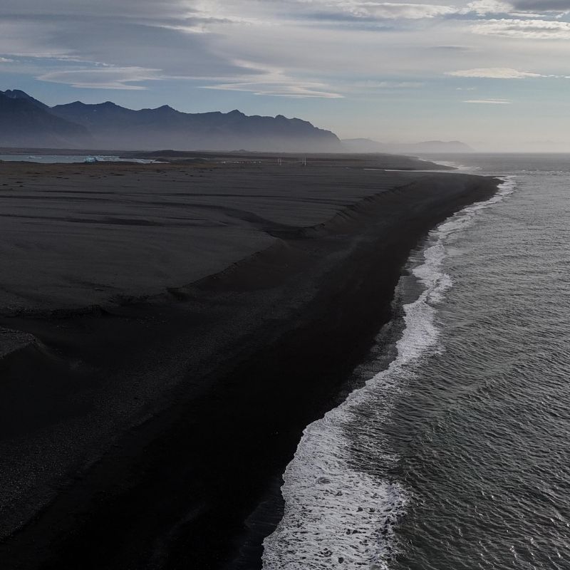 Waves meet a sweeping black sand beach on Iceland’s South Coast, with dark mountains stretching into the mist.