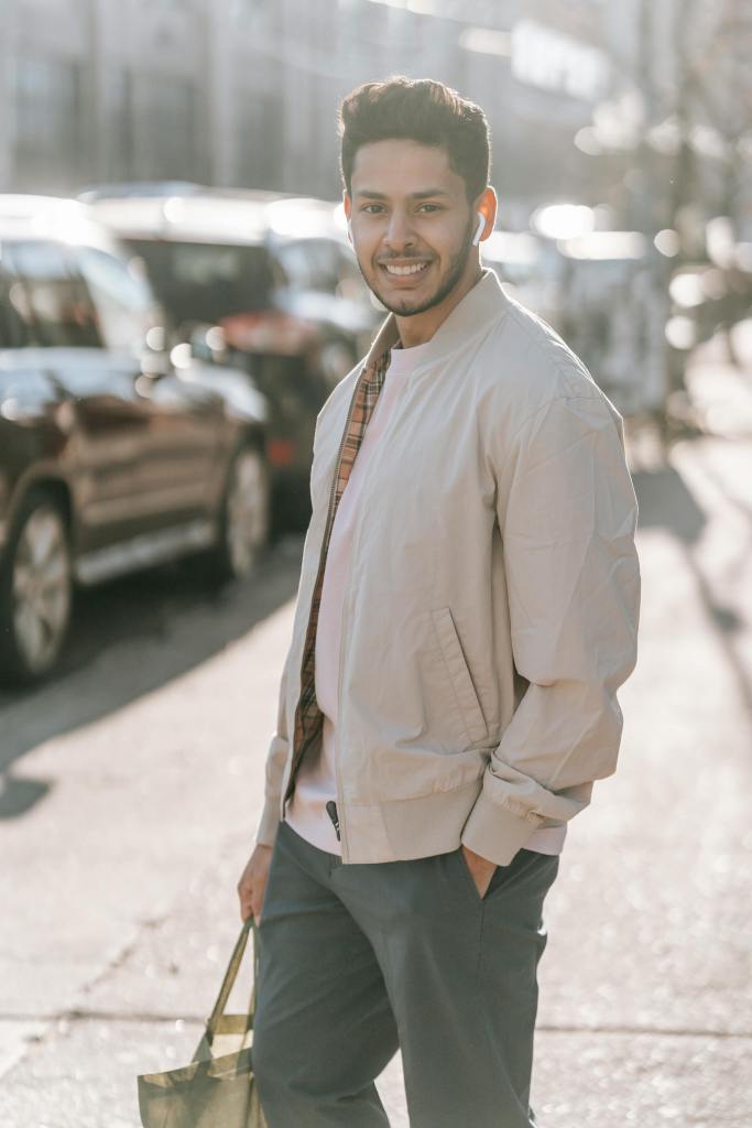 Young man with a joyful expression walking on a city street, dressed in a light jacket and carrying a tote bag, with cars parked in the background, highlighting urban life and sustainable fashion choices