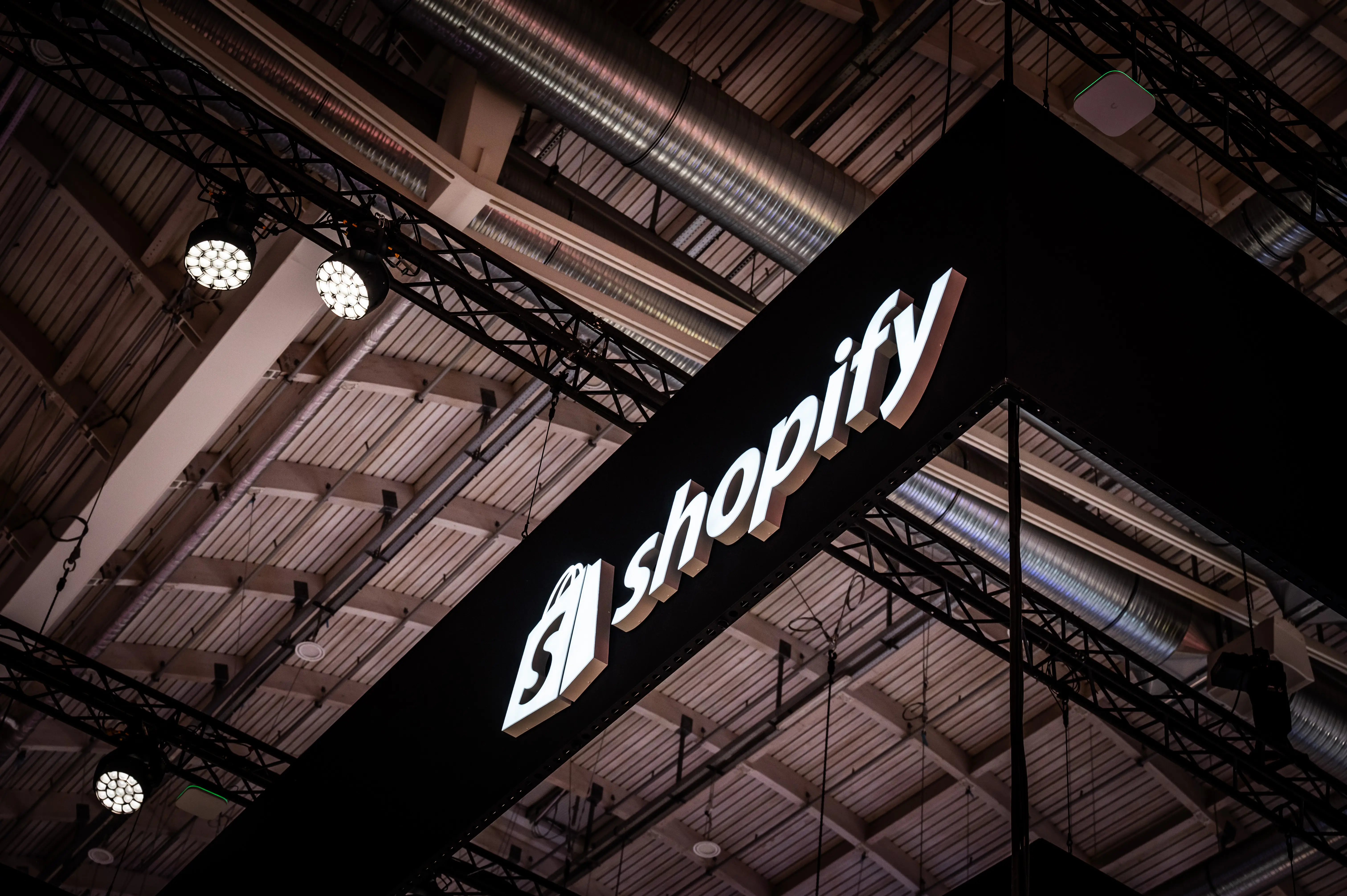 Illuminated white Shopify logo on the black hanging truss banner under the exhibition hall roof.