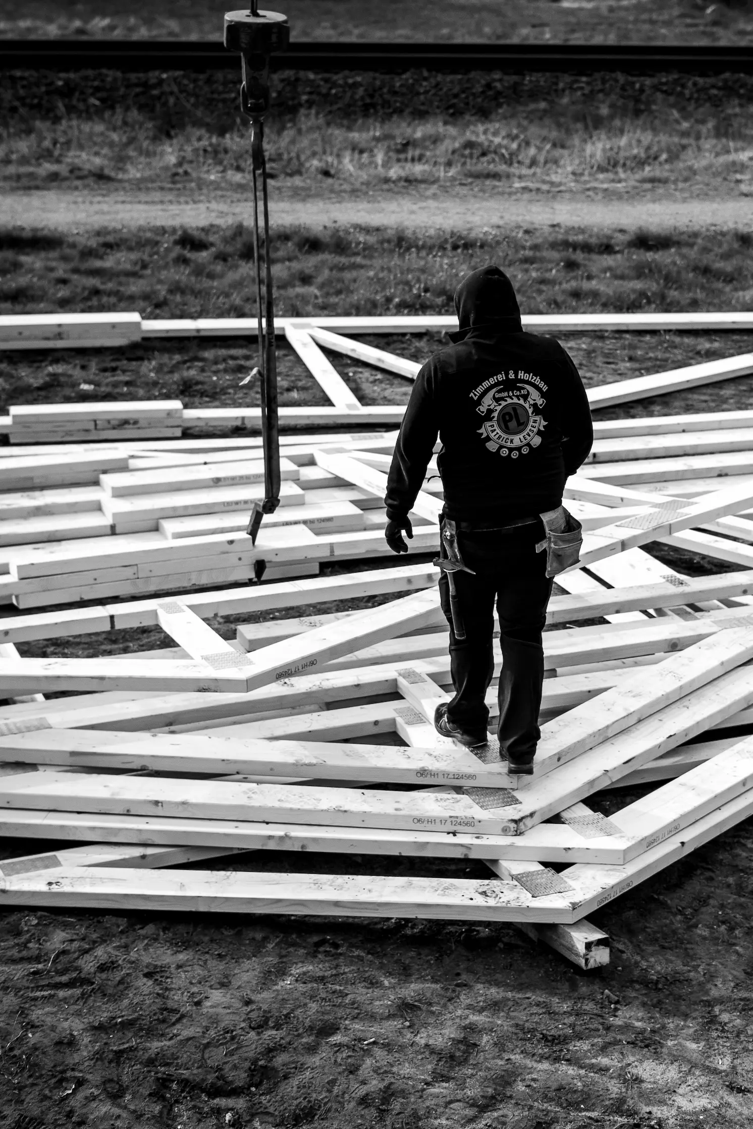 Eine Person in einem Kapuzenpullover steht inmitten verstreuter Holzbalken auf einer Baustelle; über ihr ist ein Kranhaken zu sehen, was auf Bauvorbereitungen hindeutet.