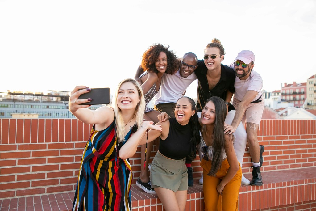 A smiling young newcomer of color looks happily at their smartphone while sitting in a modern, diverse community center or library setting in Canada, representing successful settlement and integration into Canadian culture.