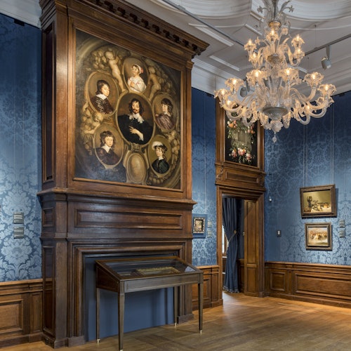 Sala de galería ornamentada con papel tapiz azul, revestimiento de madera, gran candelabro y pinturas enmarcadas, incluyendo un destacado collage de retratos.