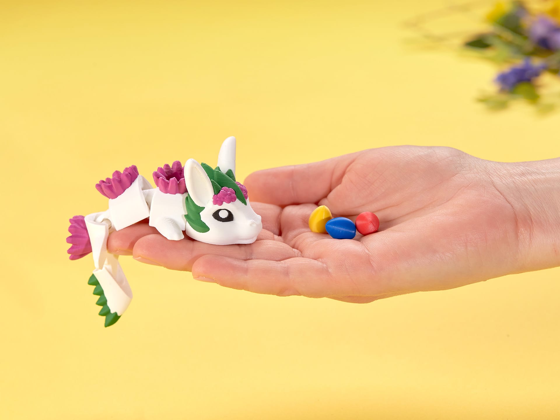 Wydrukowana w 3D biała zabawka w kształcie smoka z różowymi i zielonymi detalami spoczywa w otwartej dłoni obok trzech kolorowych jajek na żółtym tle.