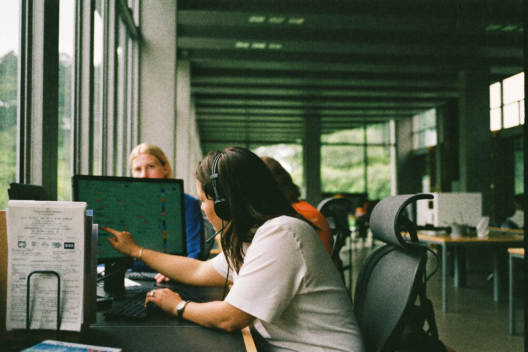 Klantenservicemedewerker wijst naar een monitor terwijl een collega meekijkt aan een bureau in een klein kantoor.
