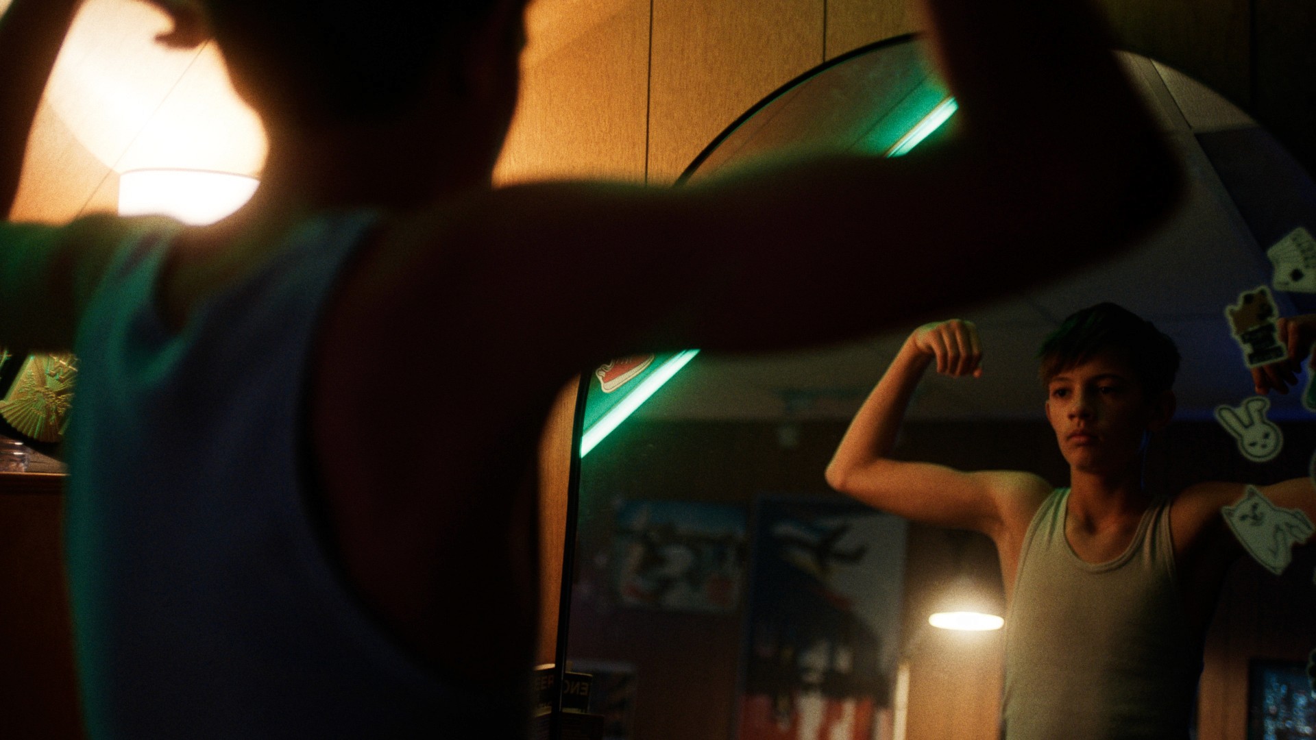 A young boy in a tank top flexes in front of a mirror, illuminated by warm light. The room has a cozy, retro vibe with stickers on the mirror.