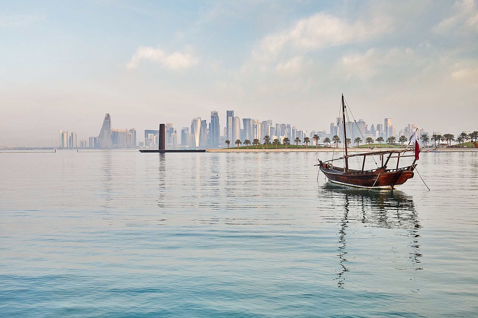 Un boutre traditionnel en bois, drapeau qatarien au mât, devant les tours de West Bay à Doha, sur une mer calme au petit matin, à l'heure où Suhail se lève