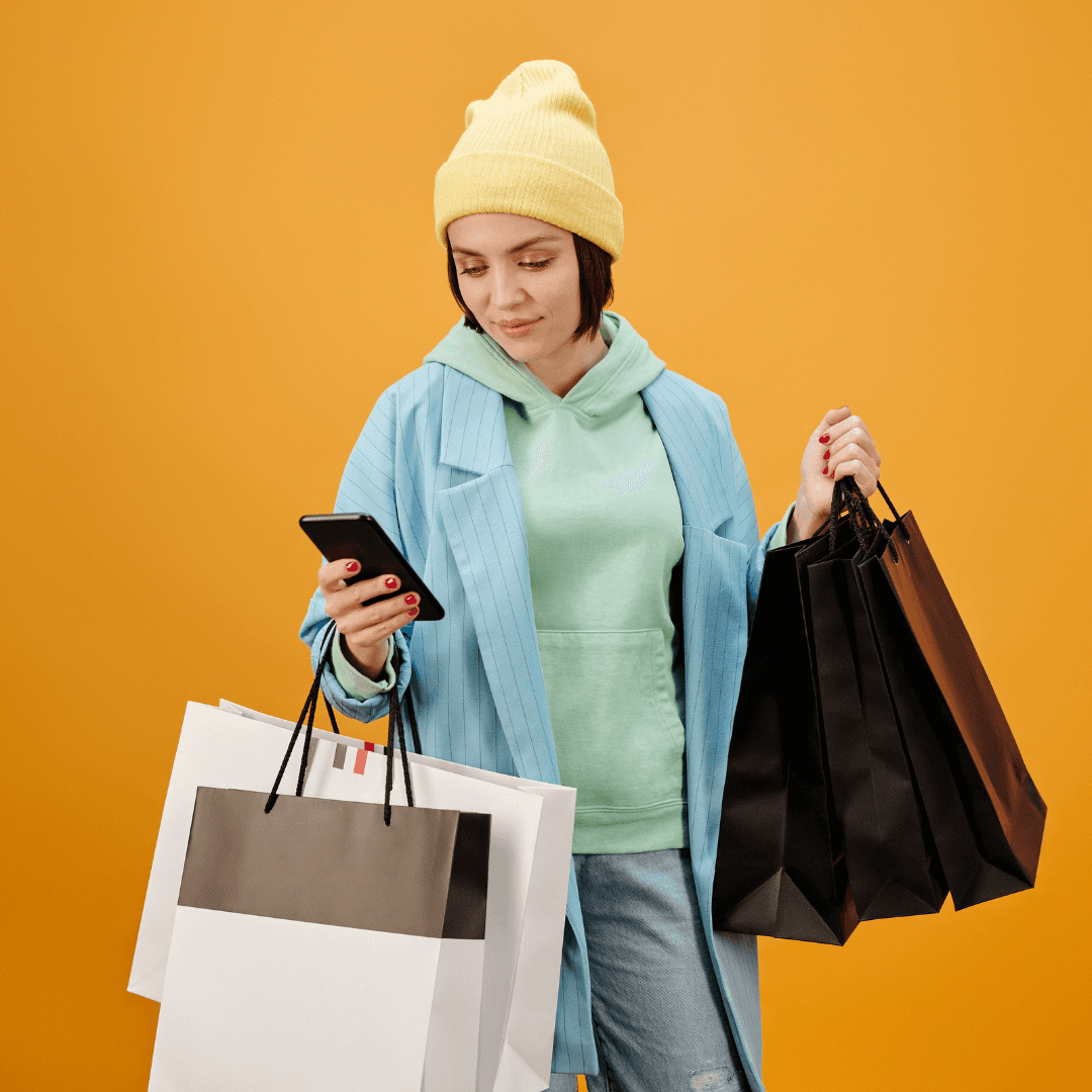 Een persoon met een gele muts en een blauw jasje houdt meerdere boodschappentassen in één hand en kijkt met de andere naar zijn smartphone, staande tegen een effen oranje achtergrond.