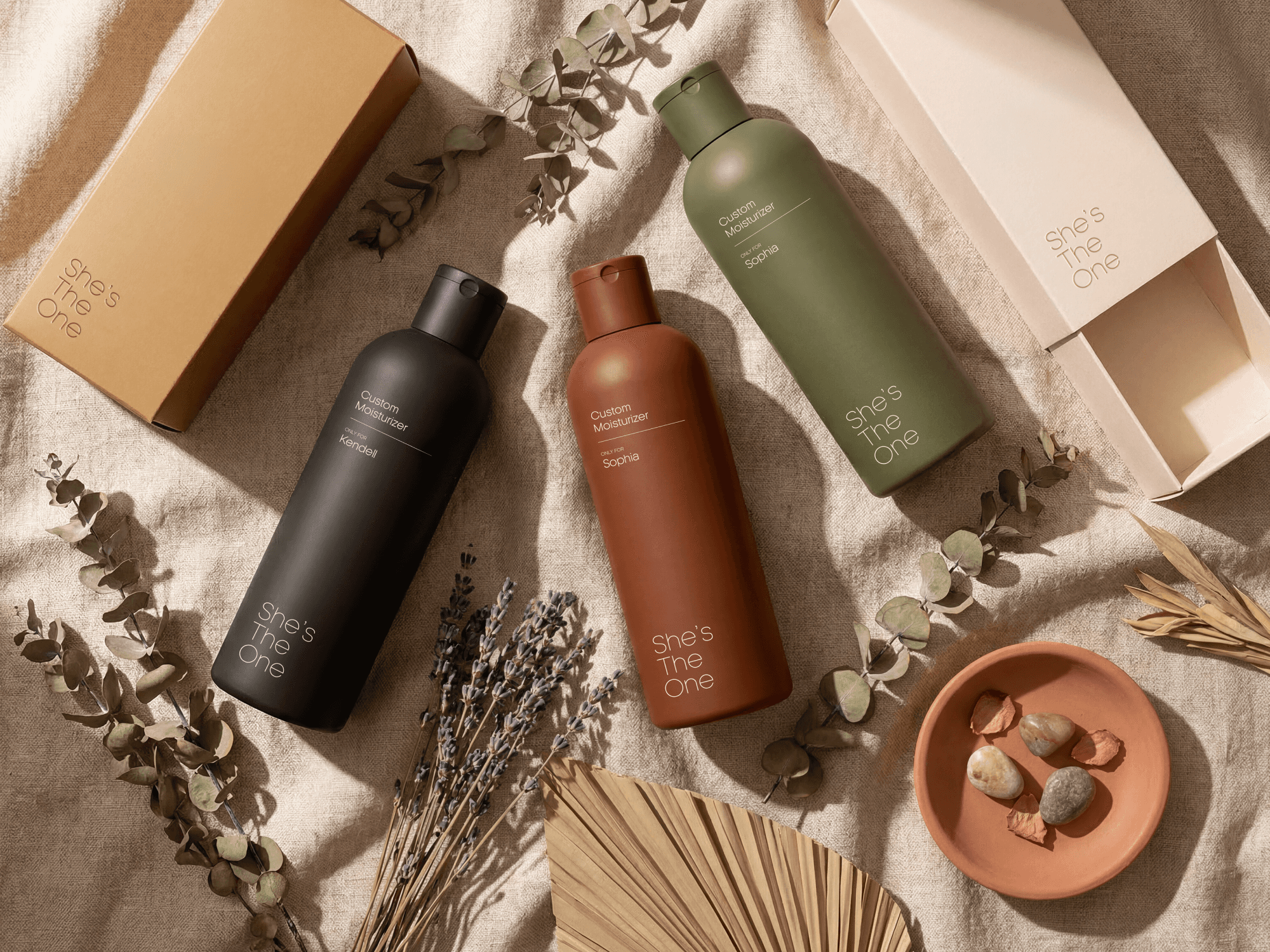 High-angle lifestyle shot of "She's The One" custom moisturizers in matte black, terracotta, and olive green bottles. The personalized packaging features names like "Sophia" and "Kendall" in minimalist white typography. The products are styled on a beige linen texture with botanical elements, dried lavender, and a terracotta dish, evoking a natural, premium, and bespoke skincare experience.
