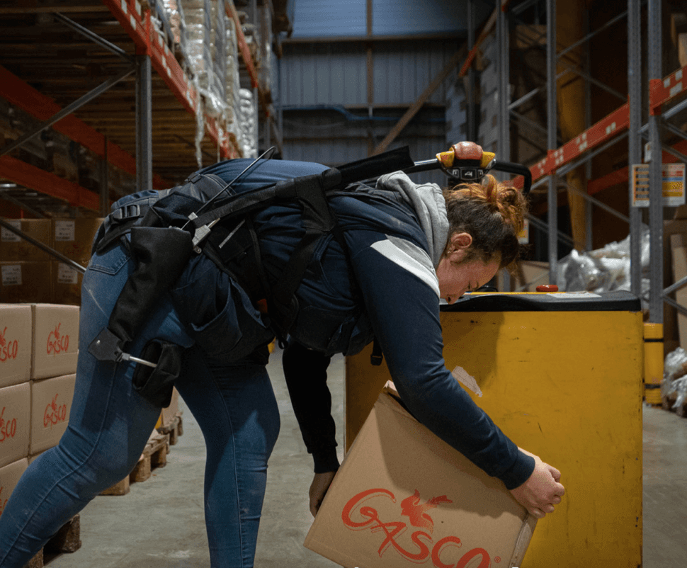 Travailleuse utilisant l’exosquelette Wave pour soulever une boîte au sol dans un environnement logistique, offrant un soutien lombaire et améliorant la sécurité au travail.