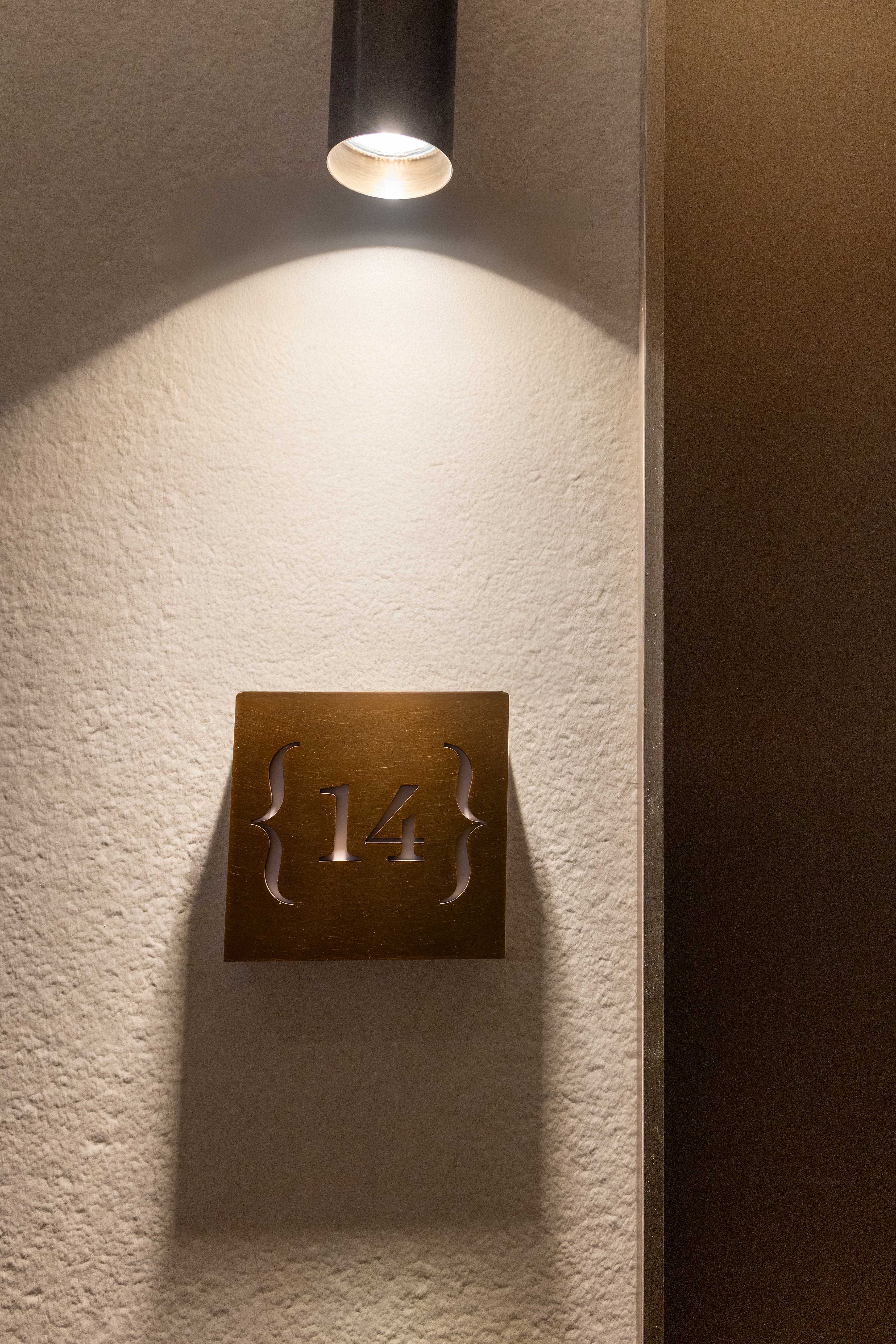 Plaque de numero 14 en laiton dore eclairee par un spot, fixee sur mur texture beige pres de la porte