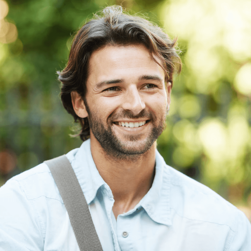 A man with medium-length brown hair and a beard smiles outdoors, wearing a light blue shirt and a shoulder strap with greenery behind him.
