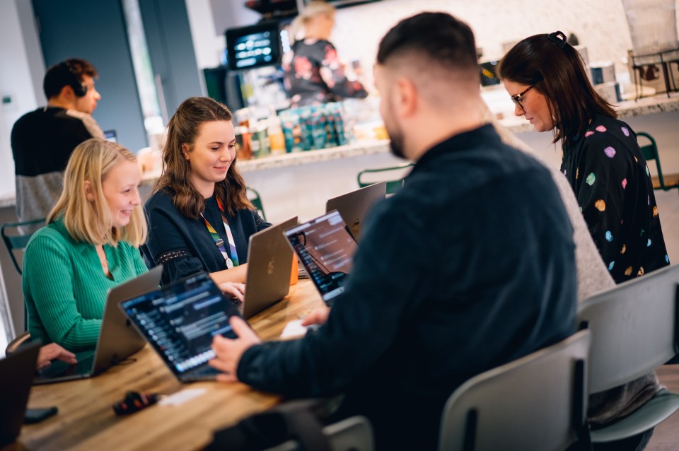 A group of people sit together around a wooden table in a bright, open workspace, each working on laptops. Several individuals appear to be discussing work while others focus on their screens. A counter and shelving area are visible in the background.