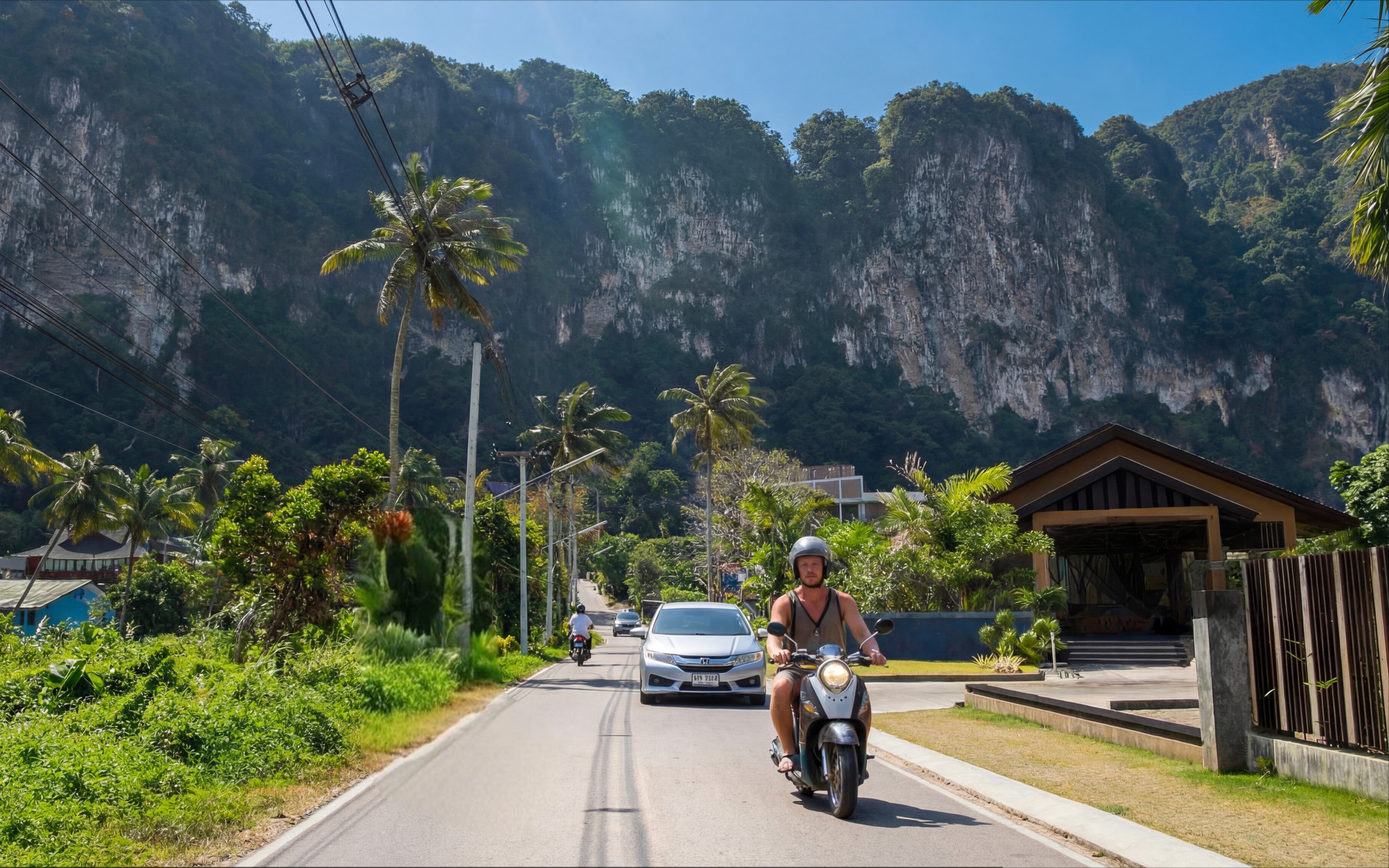 Local road in Krabi with scooters and cars passing homes and palm trees beneath limestone cliffs.