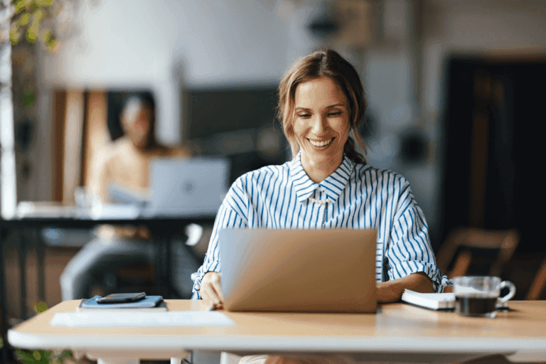 Smiling woman sitting at a desk using a laptop in a bright, modern workspace, with a notebook, phone, and coffee nearby, and a blurred coworker working in the background.