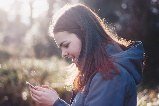 A young woman with long hair smiles while looking at her phone, surrounded by nature in soft sunlight.