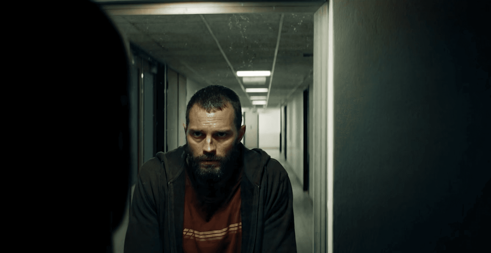 A man walks down a dimly lit hallway, his expression serious, with a blurry background.