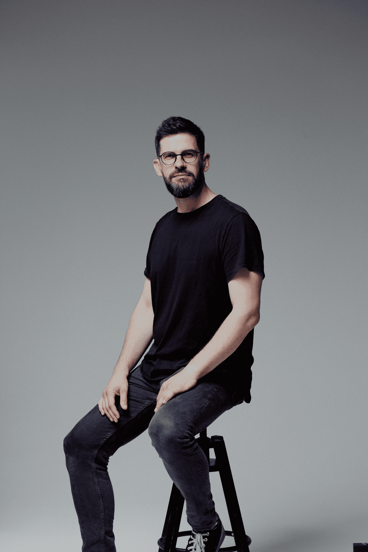 Czarno-białe półzbliżenie uśmiechniętego Bartłomieja Zylli w okularach, skomponowane centralnie na jednolitym tle