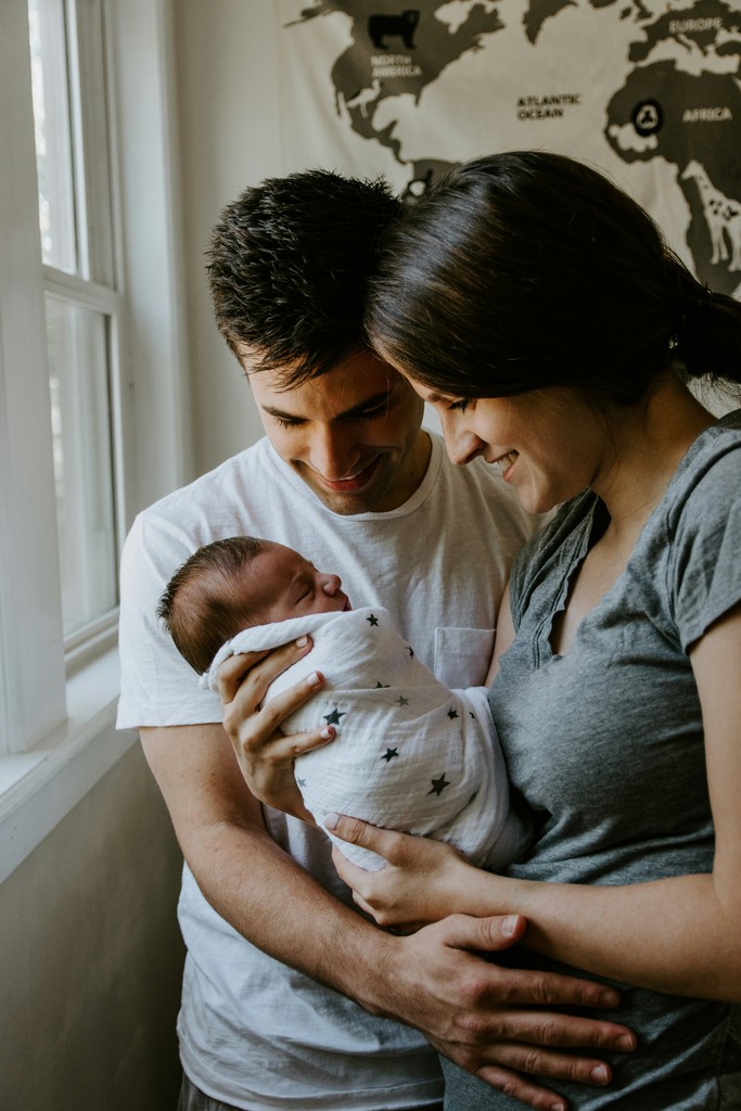 A loving couple cradling their newborn baby wrapped in a star-patterned blanket, sharing a tender moment by a window in a softly lit room, symbolizing new parenthood and family bonding.