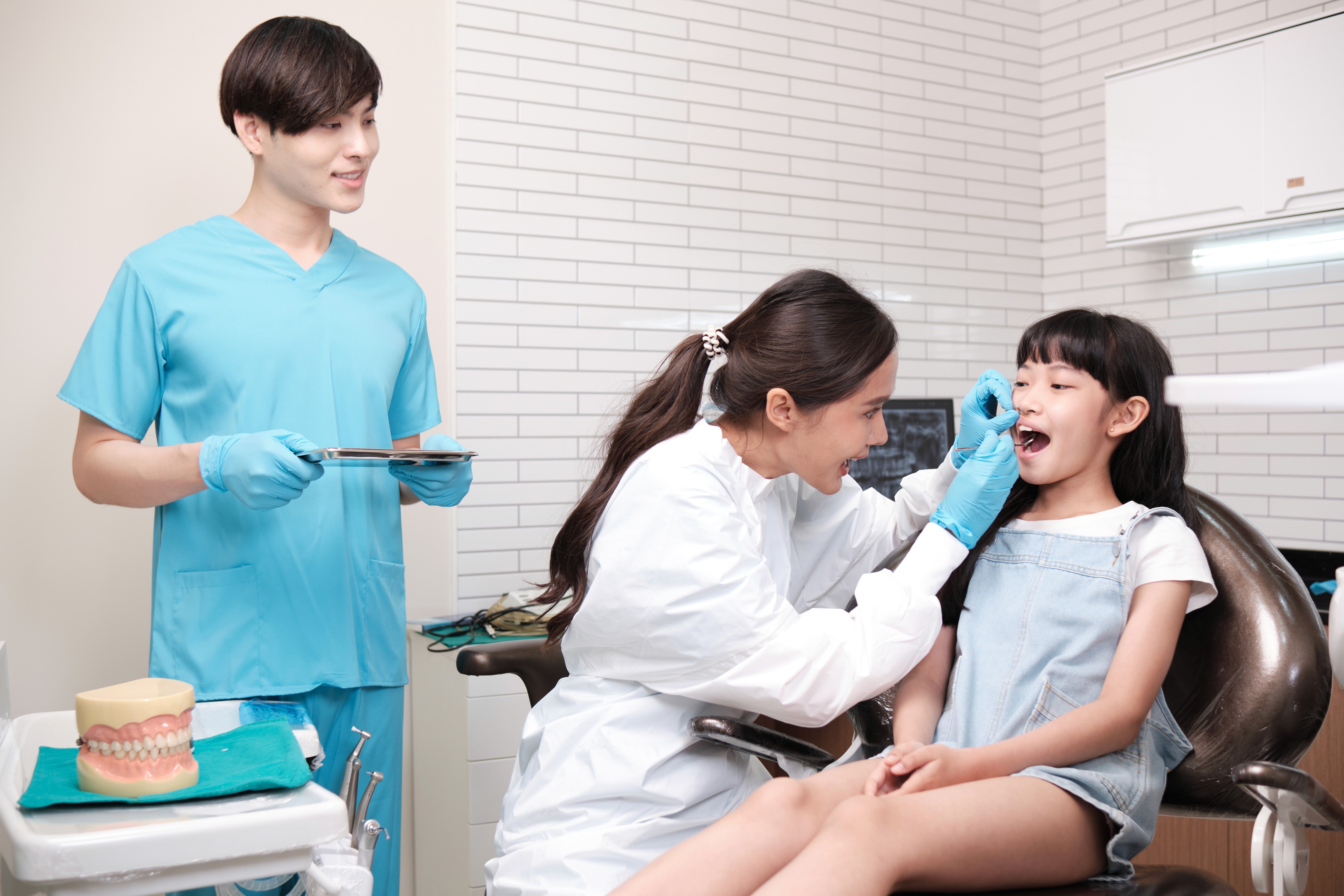 A female dentist wearing gloves examines a young girl’s teeth in a bright dental clinic. The child sits calmly with her mouth open while the dentist checks her teeth.