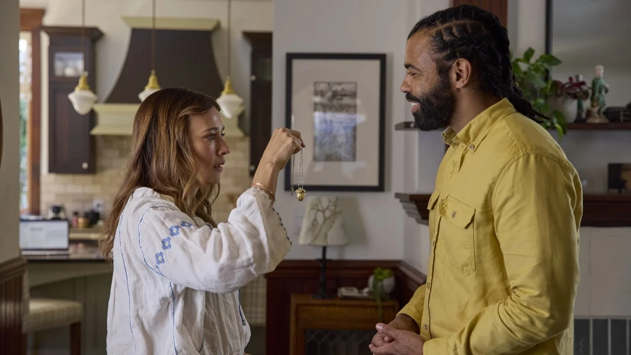 A woman in a white blouse holds up a necklace with a pendant, showing it to a man in a yellow shirt, in a warmly lit, modern kitchen with a rustic design and framed art on the wall.
