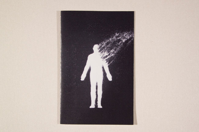 The image shows a white silhouette of a human figure standing on a black background. The figure is facing forward with its arms at its sides. A white streak extends from the figure’s head to the right side of the image. The figure and background are speckled with small white dots. The image is framed by a thin white border and is presented against a light beige or off-white background.
