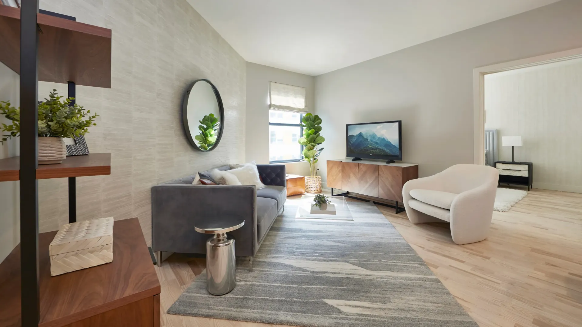 A stylish living room featuring a gray sofa, a round mirror, a TV, and modern decor, with natural light coming in.