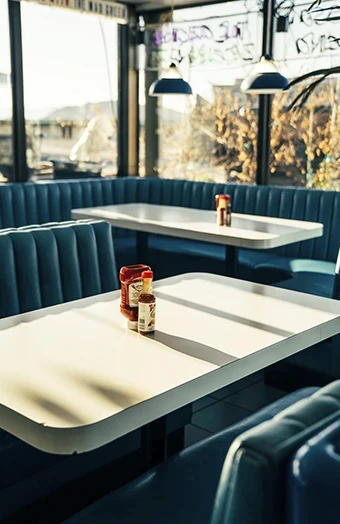 Wnętrze retro baru typu diner z niebieskimi kanapami, białymi stolikami i butelkami ketchupu oraz musztardy w świetle poranka