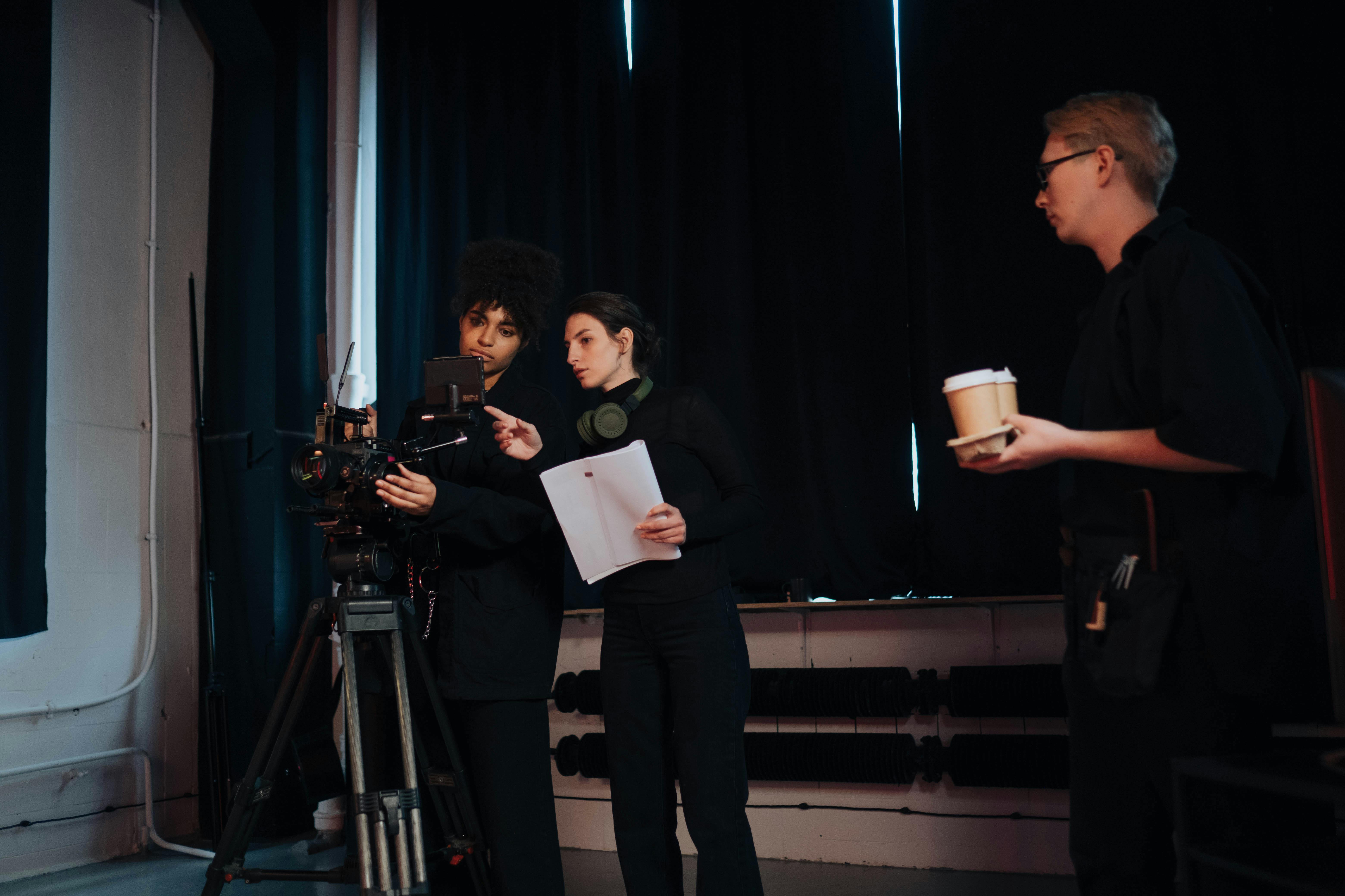 Two women review a camera monitor and script in a dark studio as a man stands nearby holding coffee.