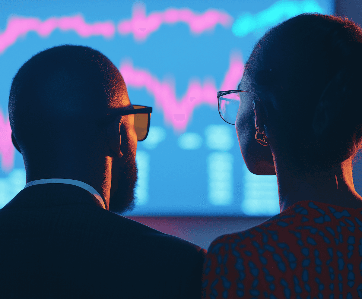 Two people viewed from behind, wearing glasses, looking at a stock market screen with colorful graphs. The setting suggests focus and analysis.