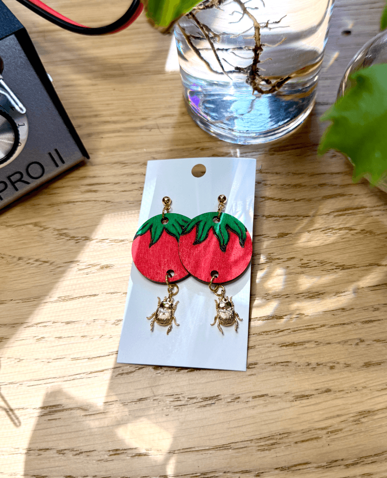 Large wood burned tomato earrings with gold plated beetle charms dangling from the bottom. The earrings are surrounded by plants, drift wood, and the colwood pro 2 wood burner.